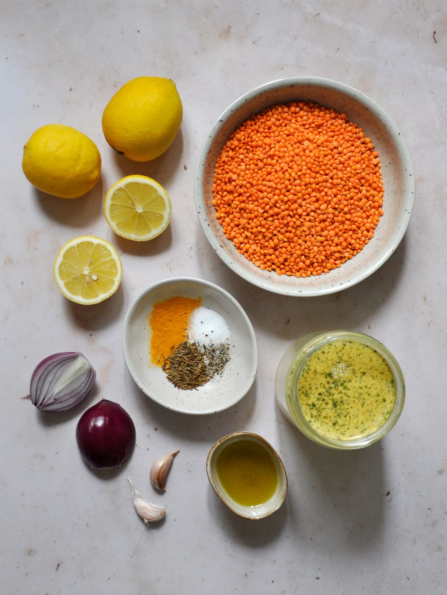 Auf dem Bild zu sehen sind die Zutaten für die vegane Zitronen-Linsen Suppe, rote Linsen, rote Zwiebeln, Knoblauch, Gewürze, Zitrone und Gemüsebrühe, auf einer Mamorplatte.
